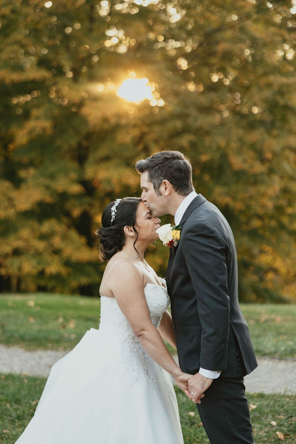 A romantic wedding couple shares a tender kiss in a sunlit autumn setting with golden foliage in the background.