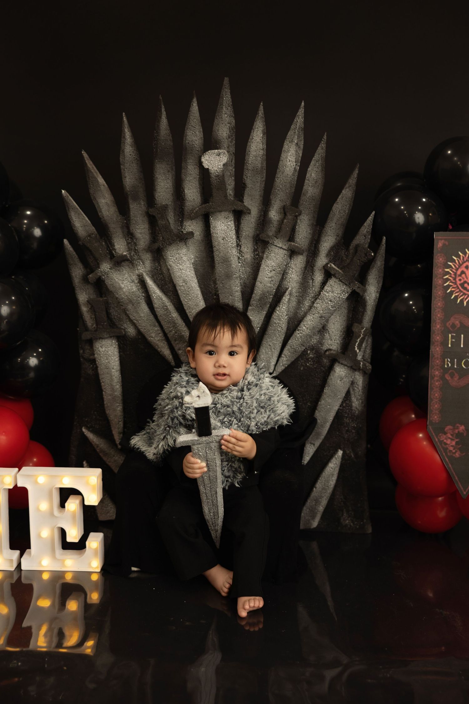 A small child sits on a replica Iron Throne from Game of Thrones with illuminated letters and red balloons in the background.
