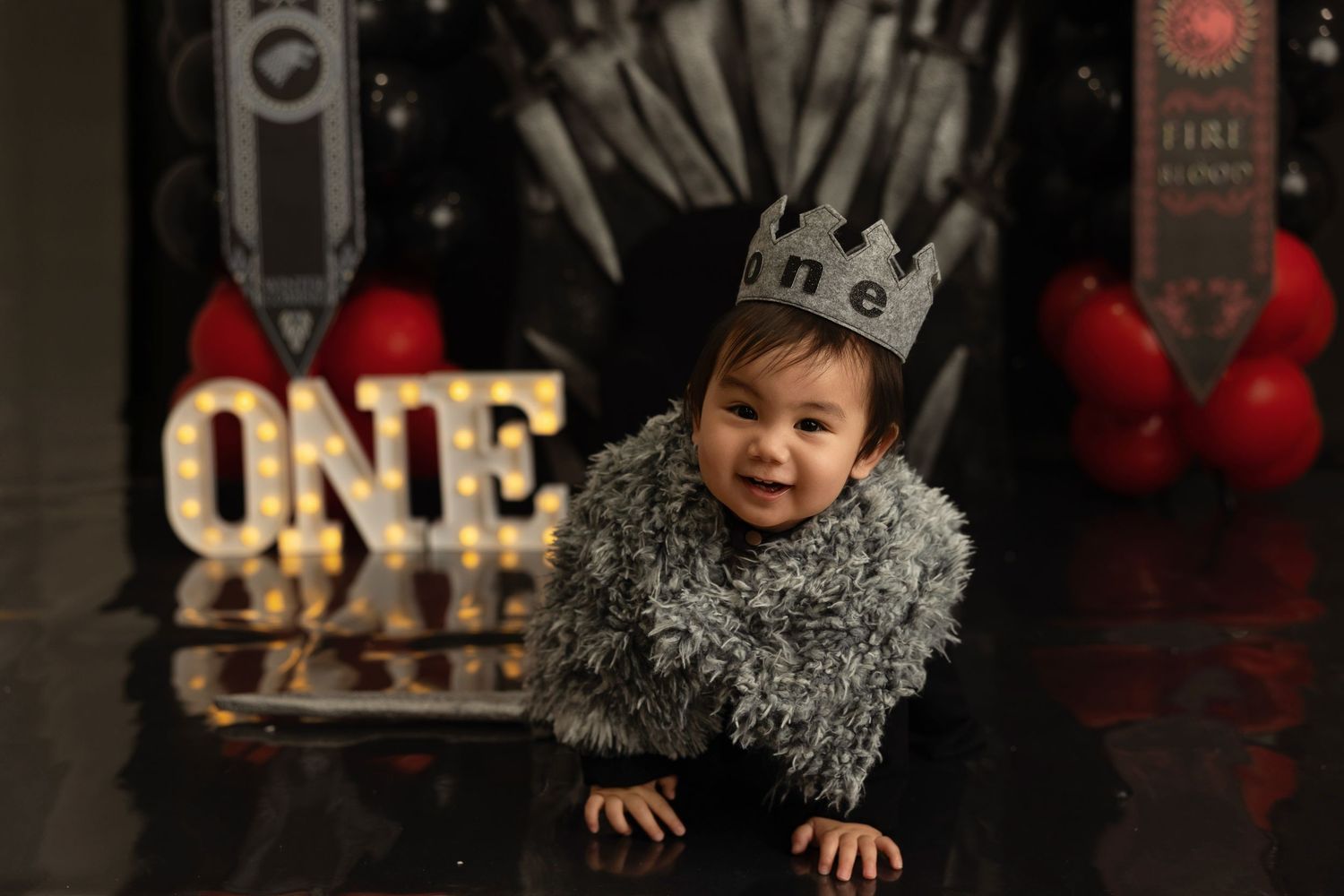 A baby wearing a sparkly silver outfit crawls on a dark floor with red balloons and party decorations in the background.