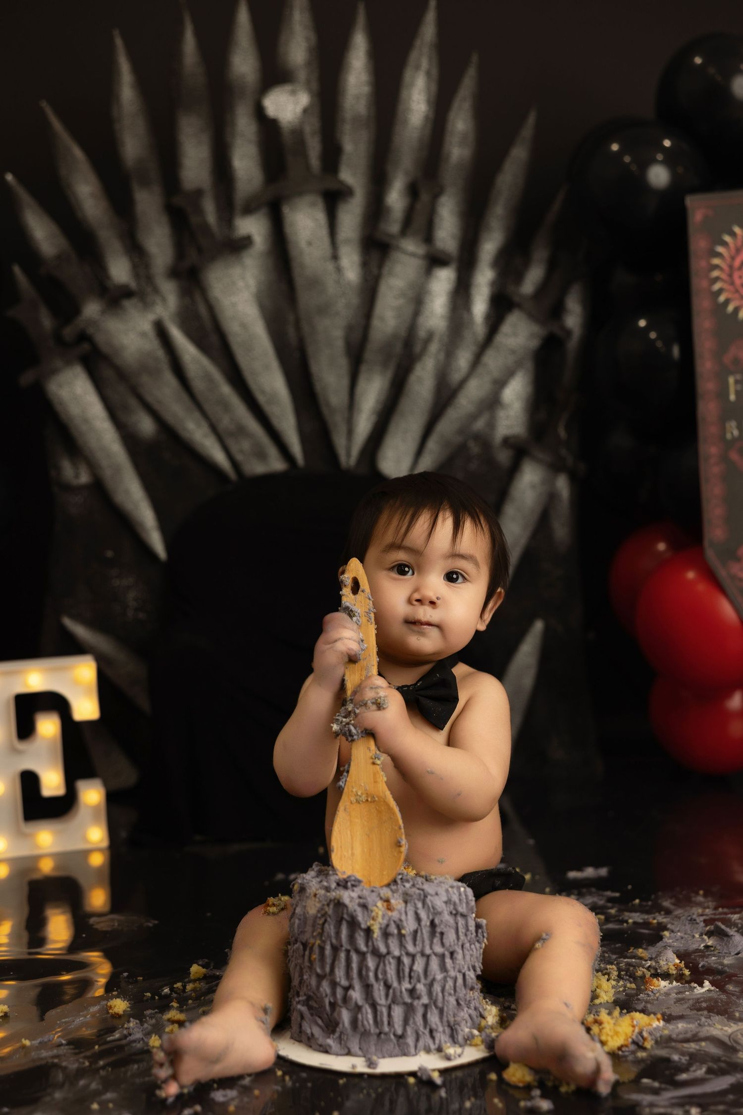 A small child sits with a cake smash setup against a dramatic throne backdrop with dark medieval fantasy styling.