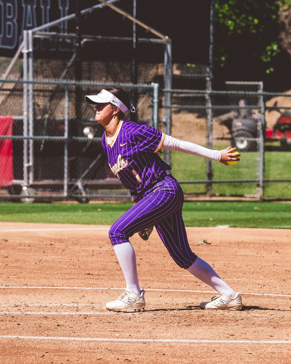Softball player in purple pinstripe uniform follows through on their pitch delivery at home plate during a game.