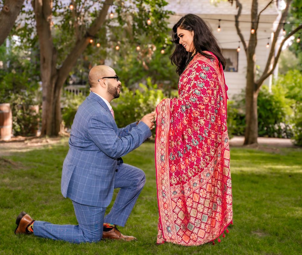 A proposal moment captured in a garden setting with someone in a blue suit kneeling before someone wearing a flowing red dress.