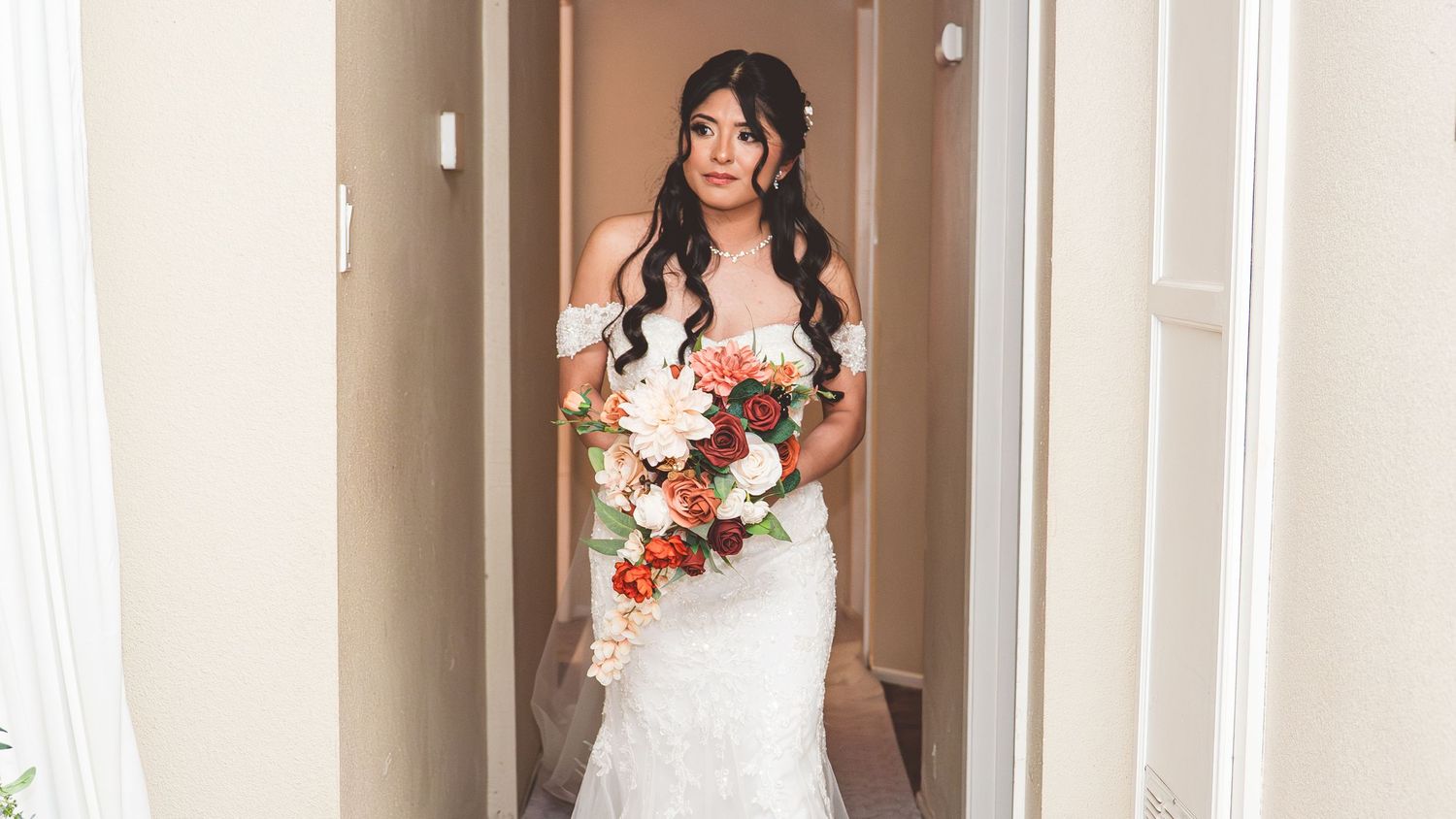 A bride in an elegant white off-shoulder wedding gown holds a vibrant bouquet of red and white flowers in a hallway.