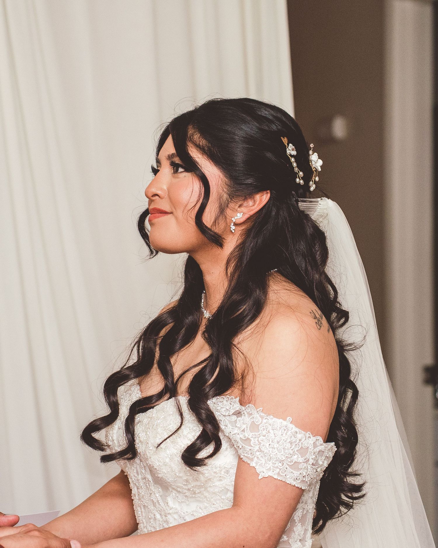 Bride in an off-shoulder lace wedding dress smiles while looking to the side, with long dark curled hair and delicate hair accessories.