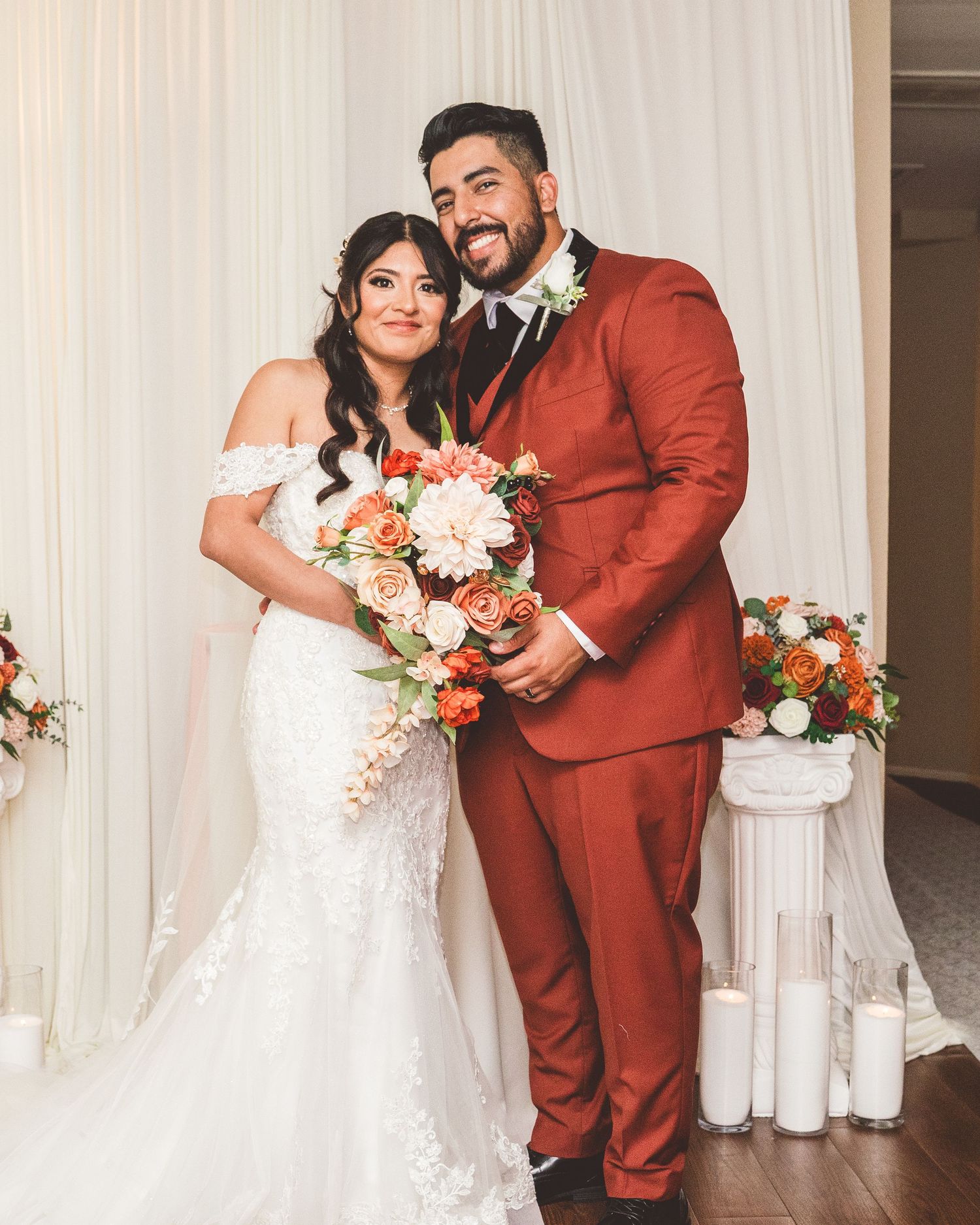 A couple in a white wedding dress and rust-colored suit embrace and laugh together during their wedding celebration.