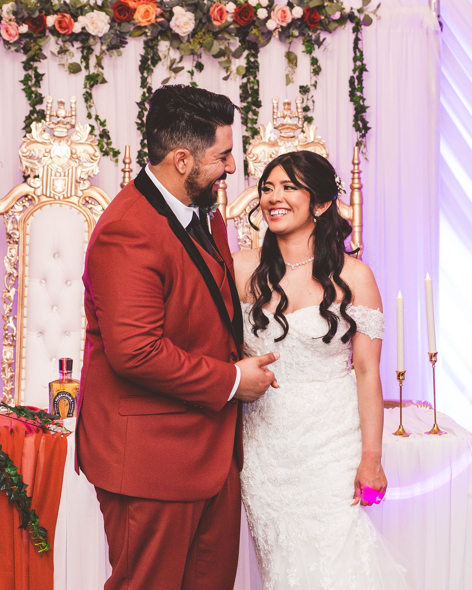 A couple shares a joyful moment at their wedding under an ornate floral arch with pink and coral roses.