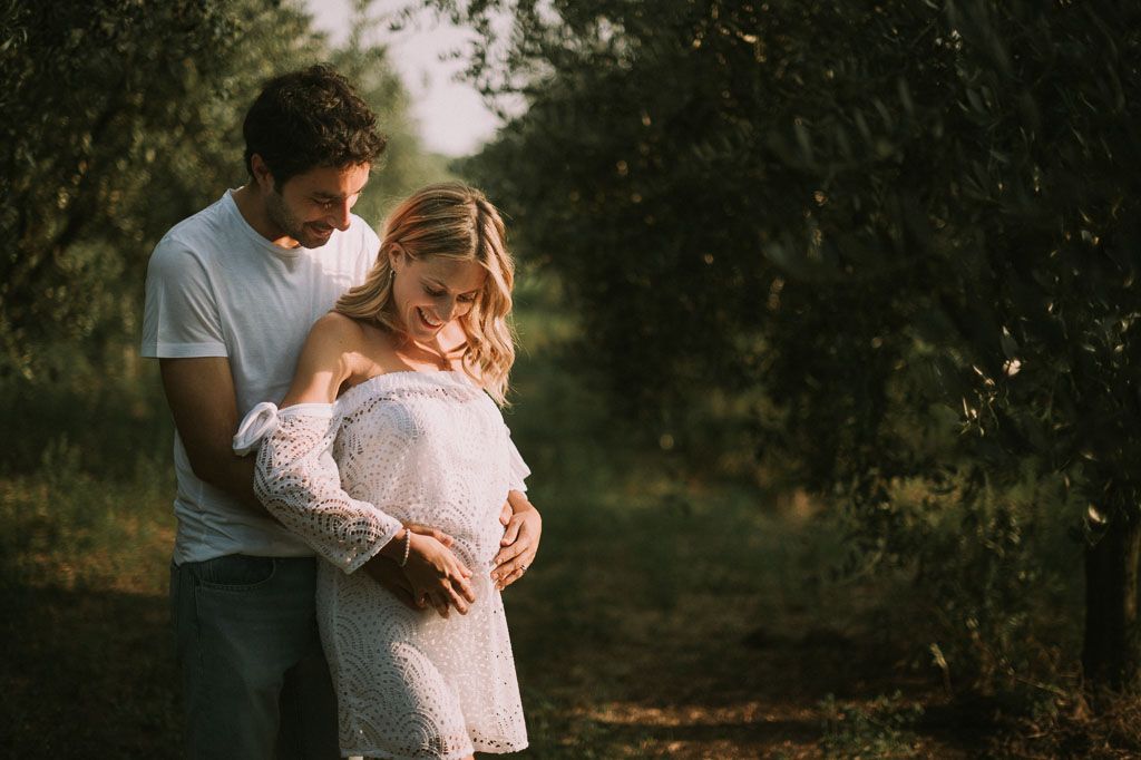 A romantic couple embraces in a sunlit orchard during a golden hour maternity photoshoot.