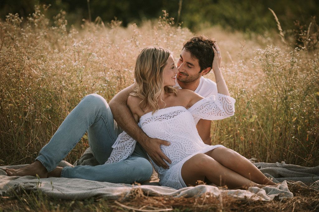 Couple lounges on blanket in golden field during sunset maternity session.