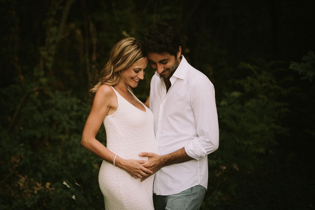 Maternity photoshoot in dark moody setting with couple in white dress and casual wear.