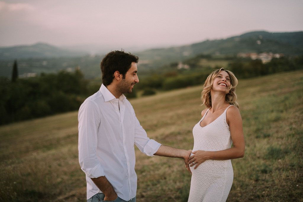 A couple in white and casual attire walks hand in hand through a scenic mountain landscape at dusk.