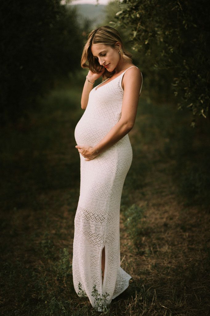Expectant mother in white lace dress poses in natural outdoor setting.
