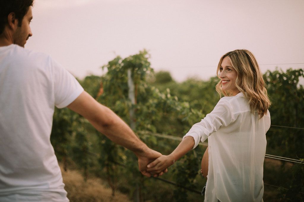 A couple in white clothing holds hands while walking through a scenic vineyard at sunset.