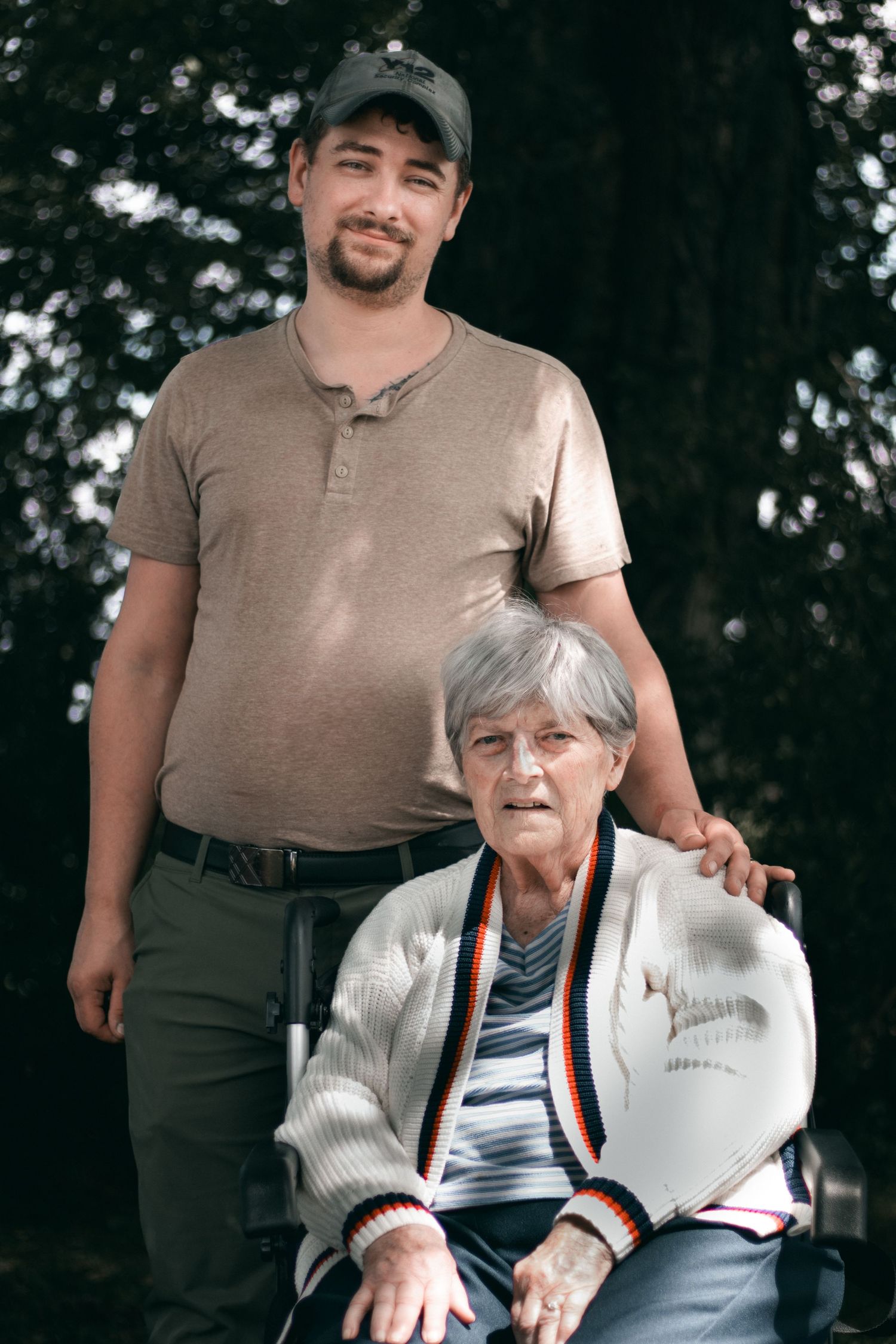 A person in a beige shirt stands behind someone in a white sweater sitting outdoors.