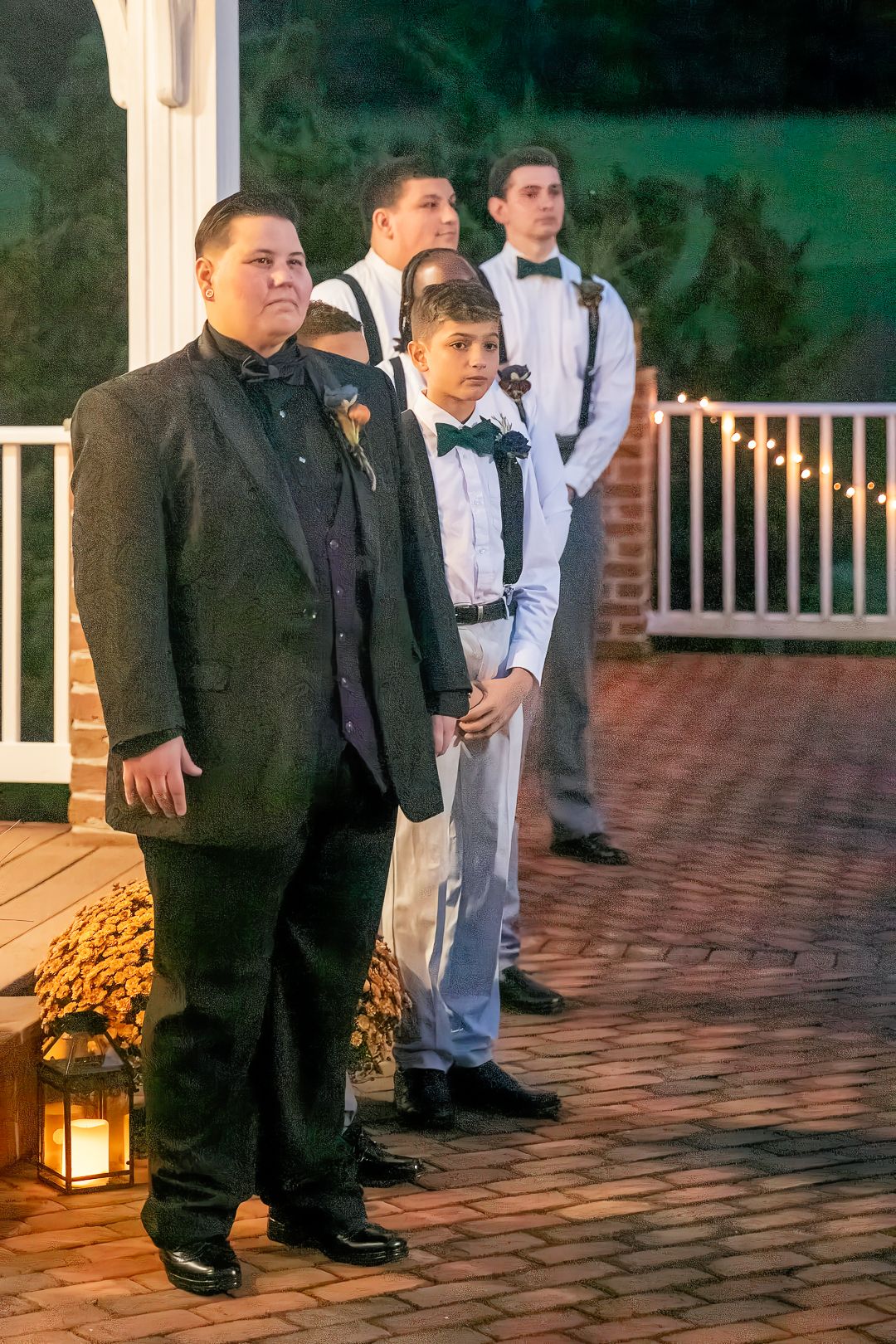 Groomsmen in black suits and light colored ties stand on brick patio at evening wedding reception with lantern decor.