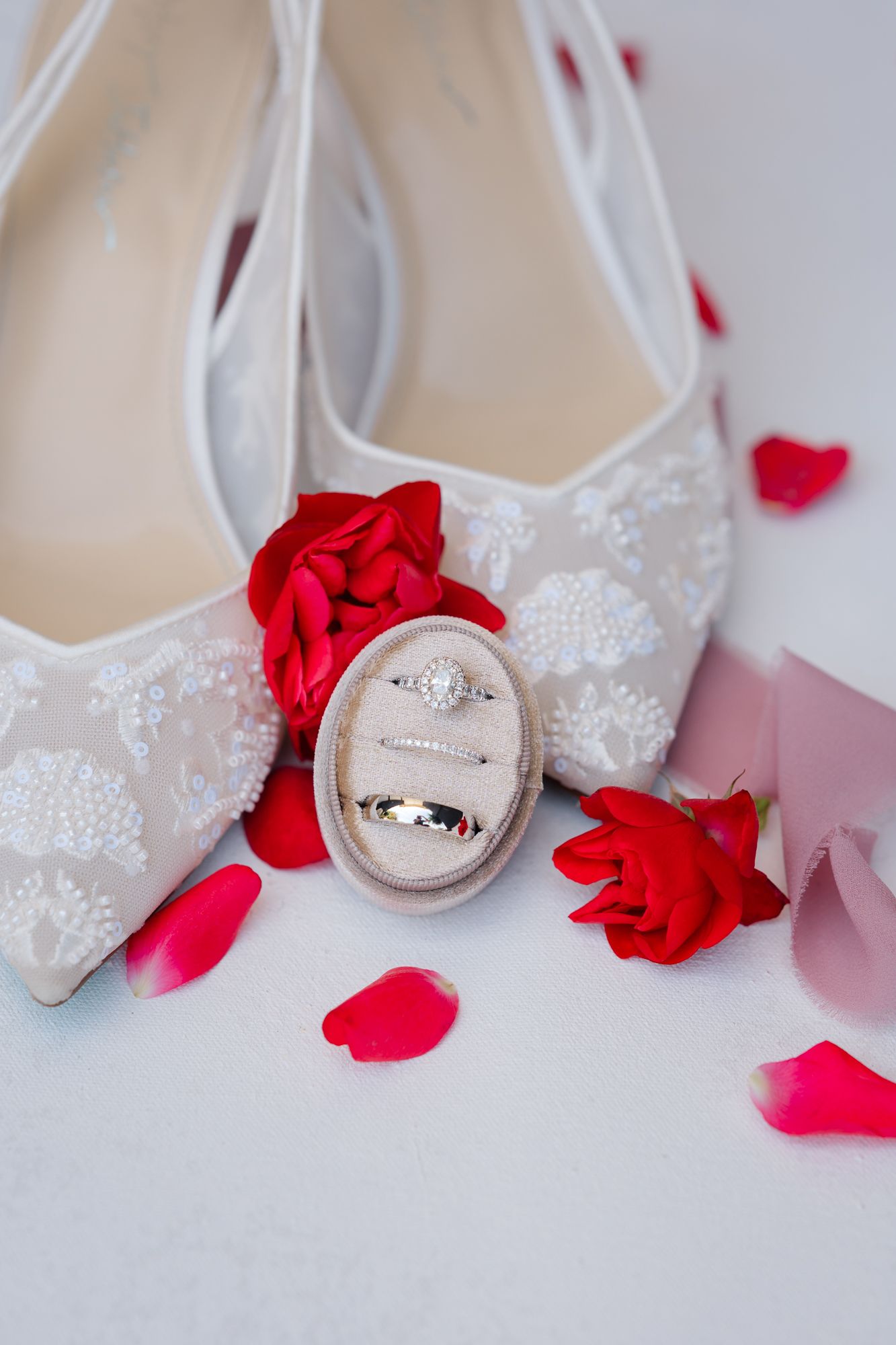 White lace wedding shoes with red rose petals and wedding rings arranged on a light background.