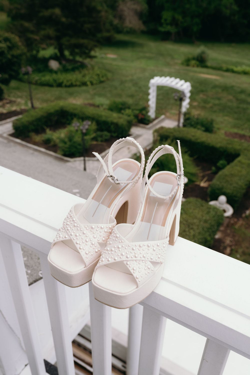 Delicate white lace bridal shoes displayed on a white railing with green landscape in background.