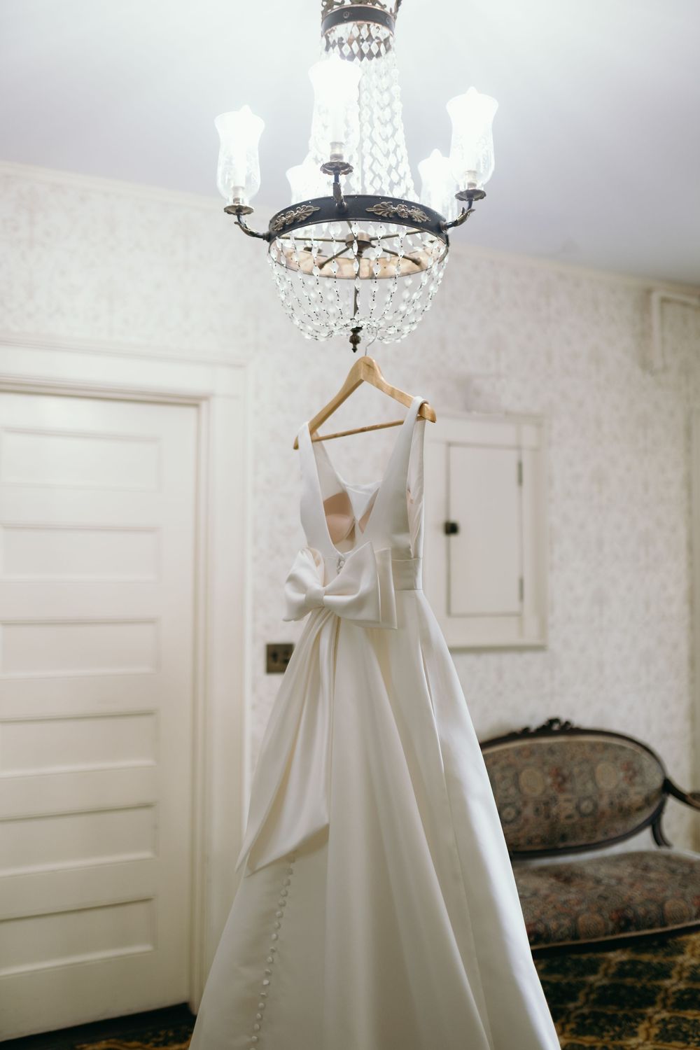 Elegant white wedding dress hanging from chandelier in bridal suite with classic decor.