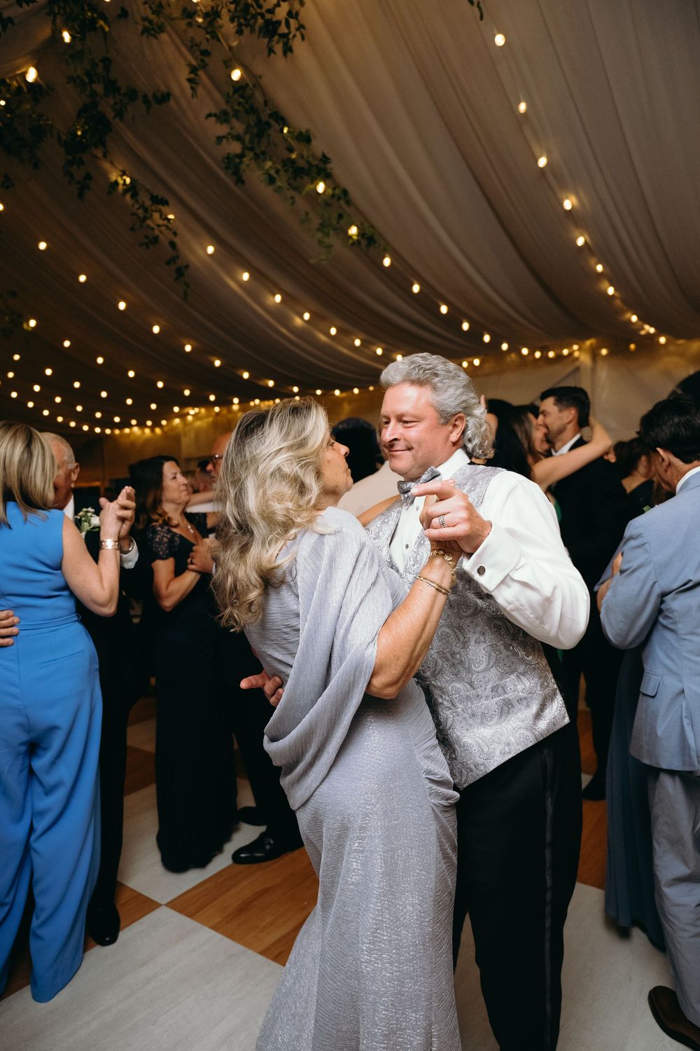 Couples dancing together under string lights at evening wedding reception in tent.