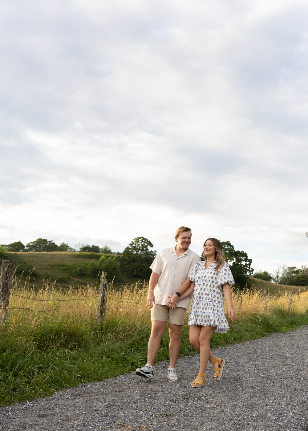 Couple walking on a gravel path through green fields, woman in floral dress and man in beige outfit.