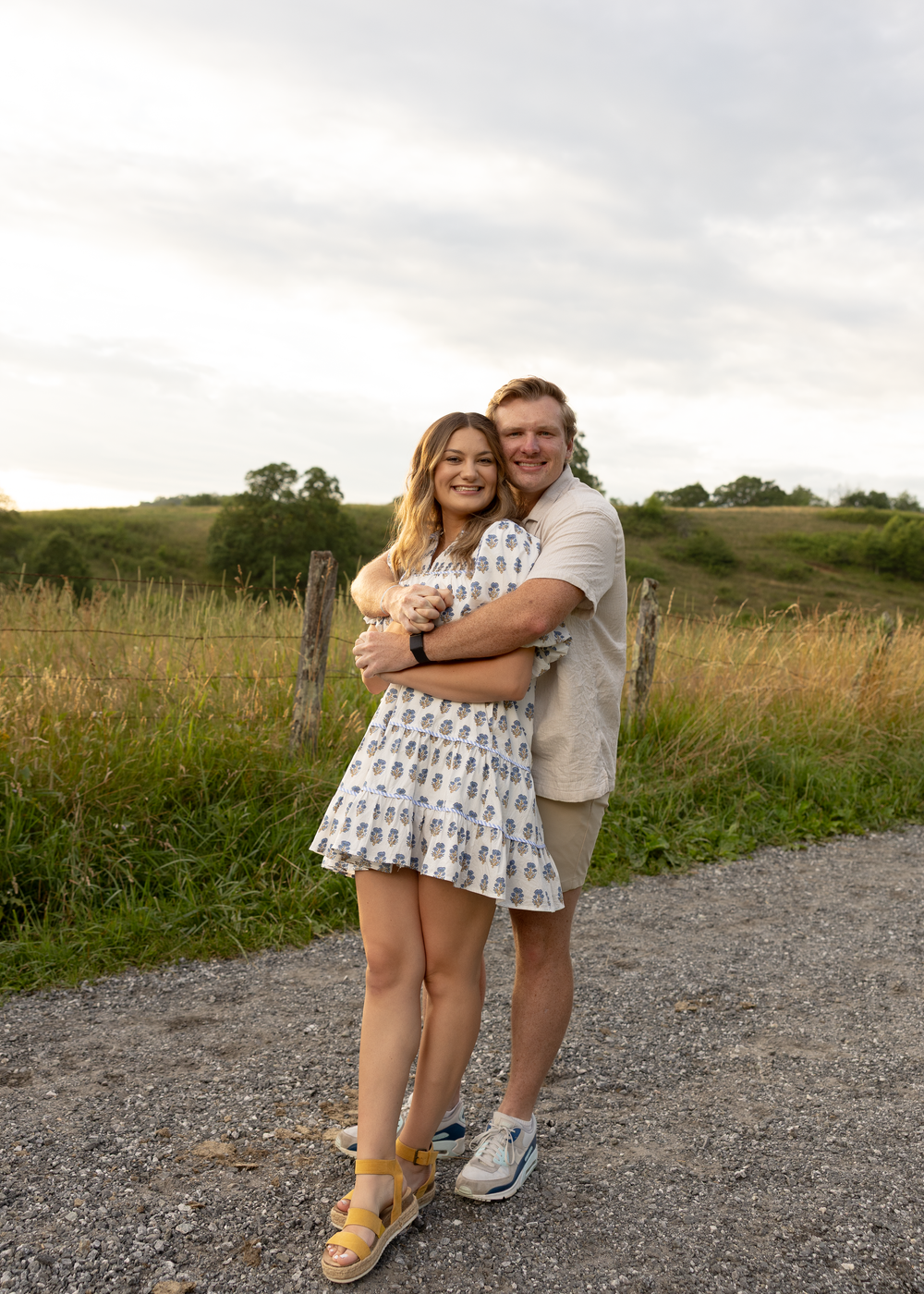 Happy couple hugging on a gravel path with rolling green hills and fence in the background.