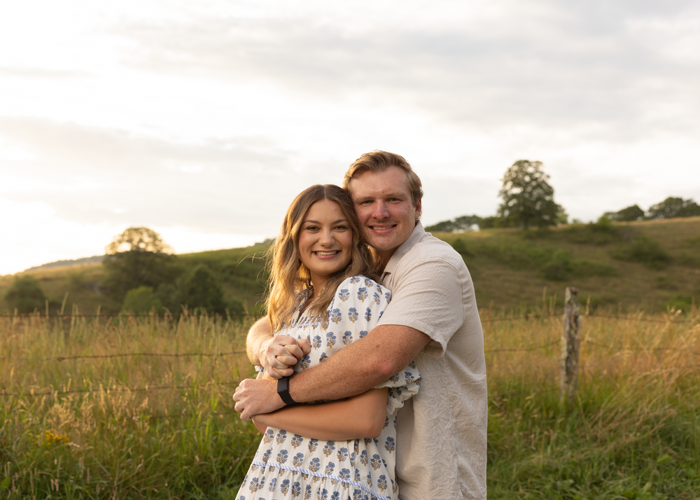 Smiling couple embracing in a golden meadow at sunset, woman in floral dress and man in white shirt.