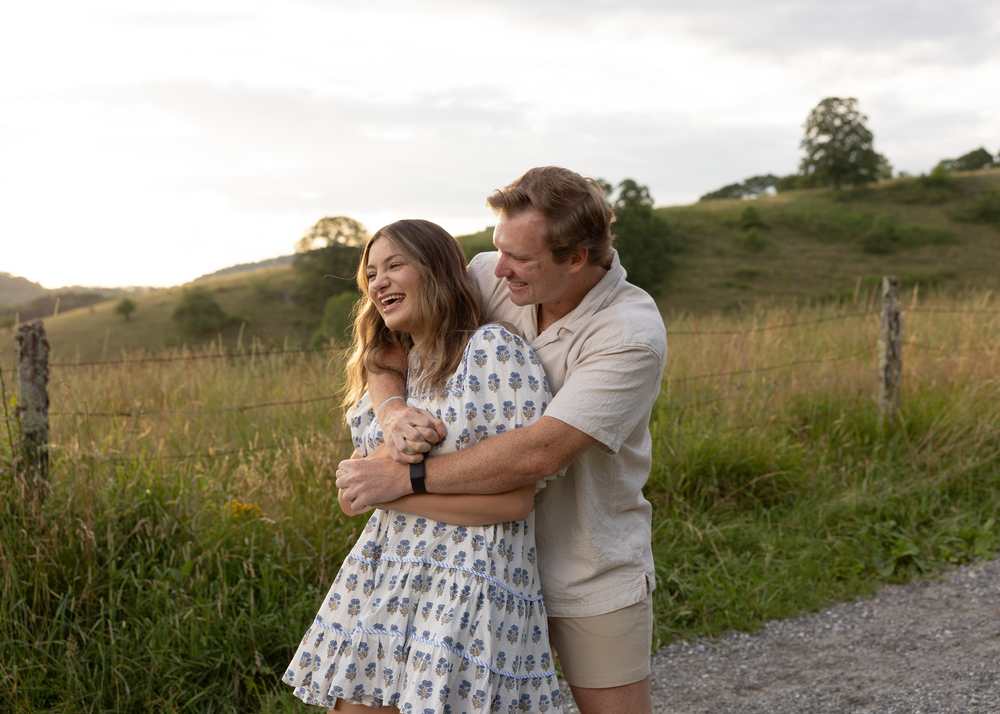 Joyful couple laughing together while embracing on a scenic gravel path surrounded by green fields.