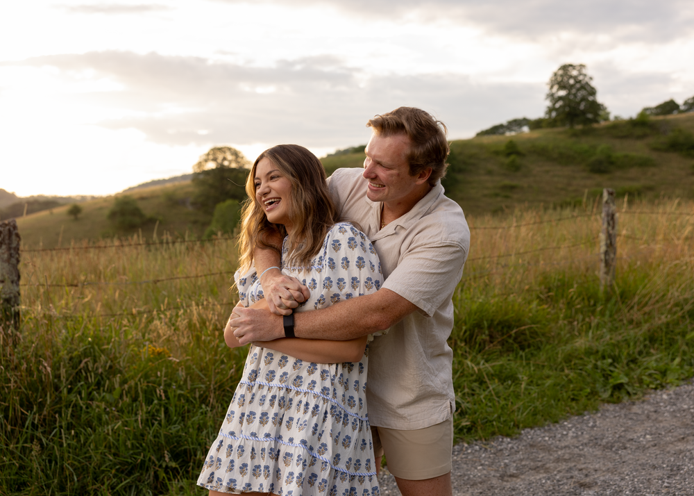Couple sharing a laugh while embracing on a countryside path with green hills in the background.