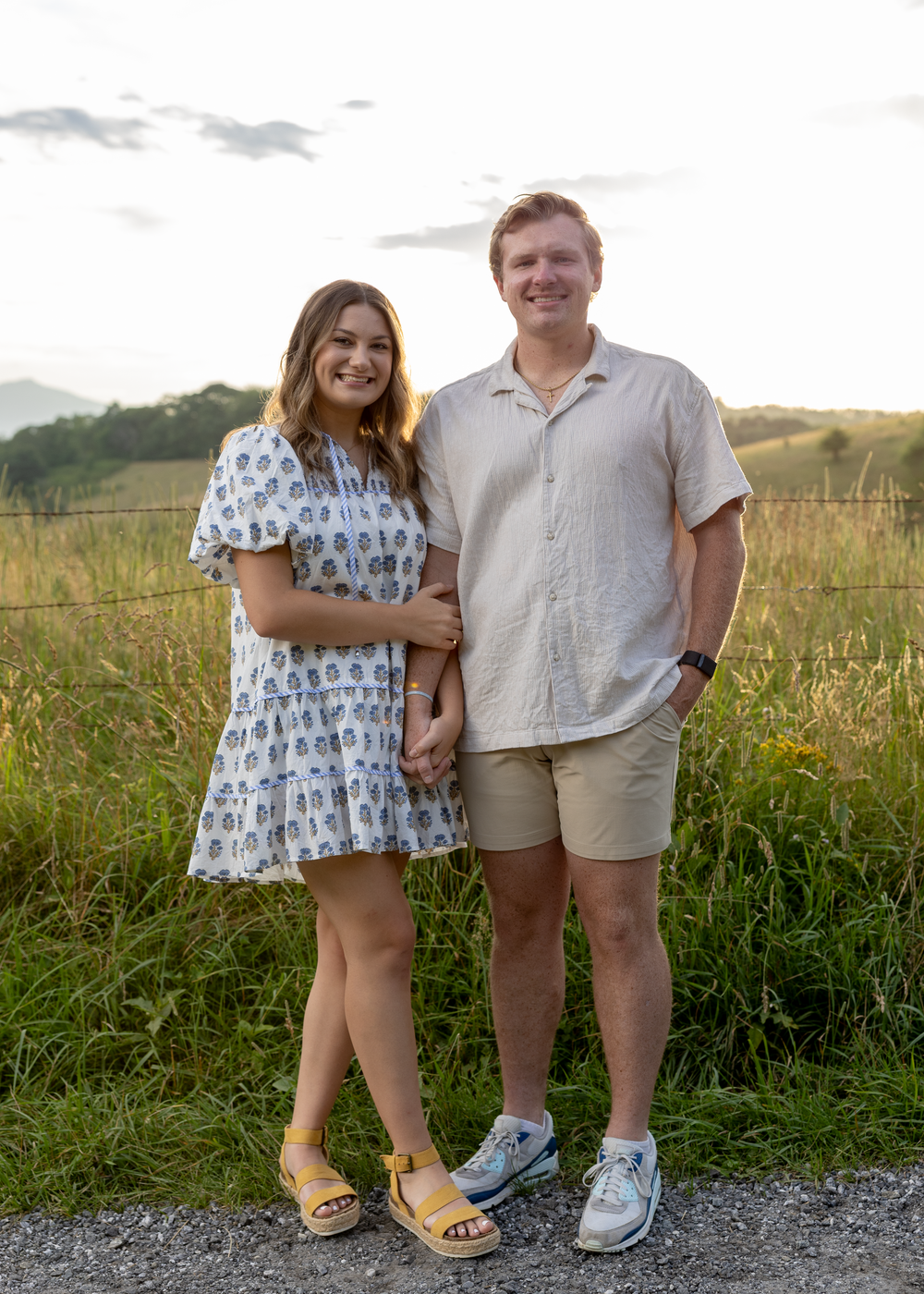 Smiling couple holding hands on a gravel path with golden meadows and hills behind them.