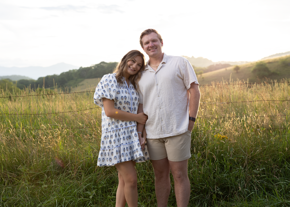 Couple posing together in a sunlit meadow, woman in floral dress and man in beige shirt and shorts.