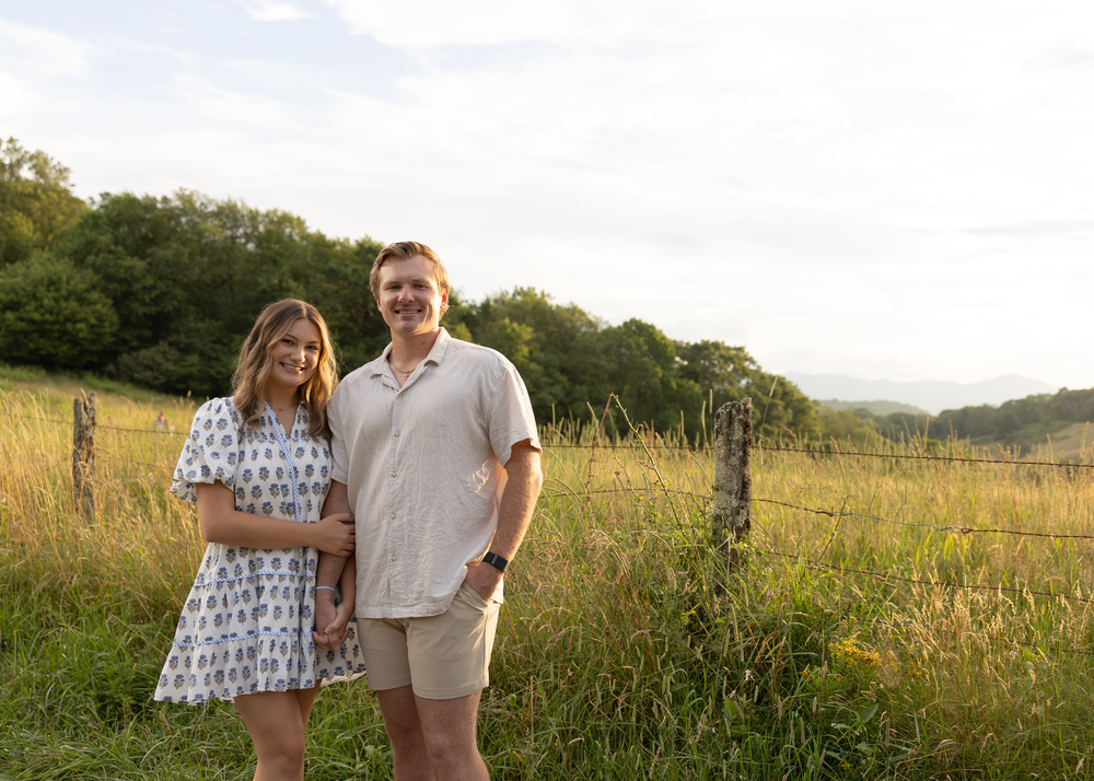 Couple smiling together in a sunny field with rolling green hills and a rustic fence in the background.
