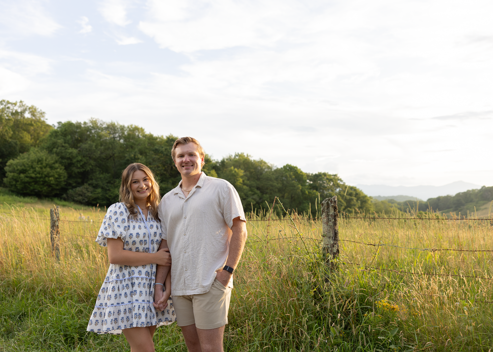 Happy couple posing in a lush meadow with tall grass, trees, and a cloudy sky behind them.