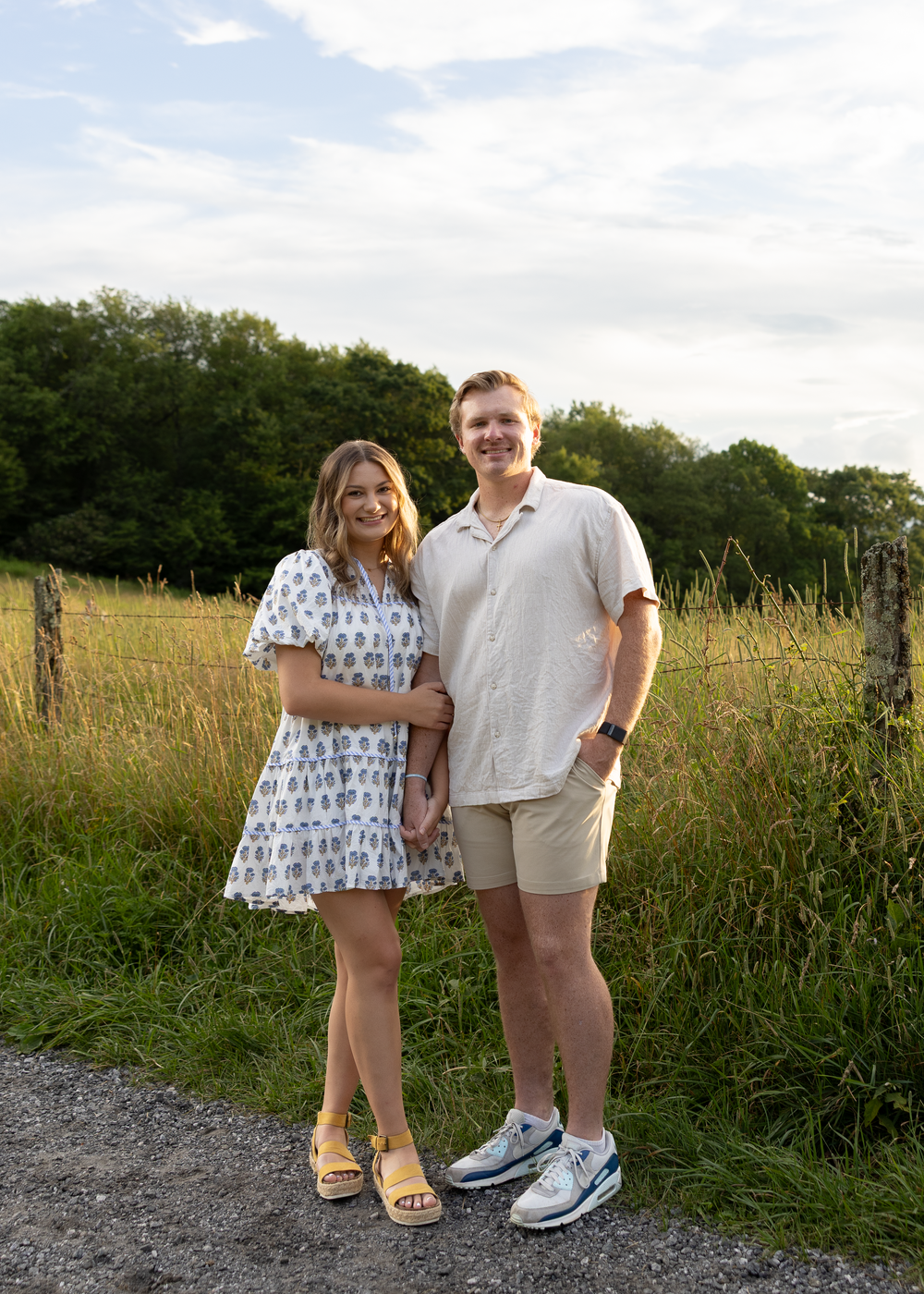 Smiling couple standing on a gravel path beside a green field, woman in floral dress and man in khaki shorts.
