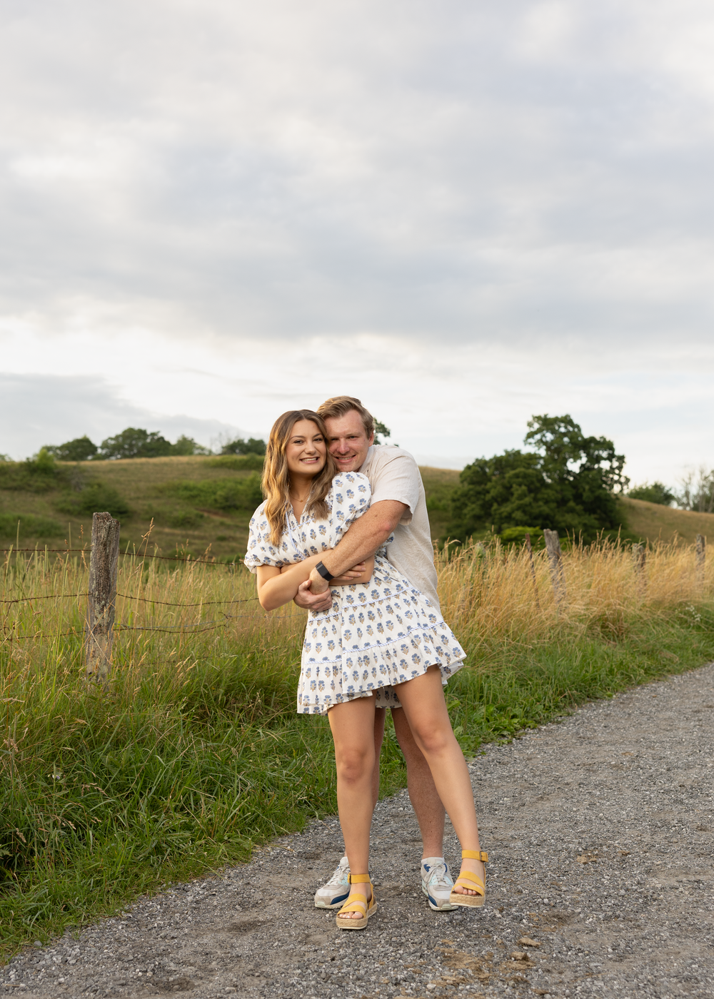Joyful couple hugging and laughing on a scenic gravel path surrounded by green countryside.