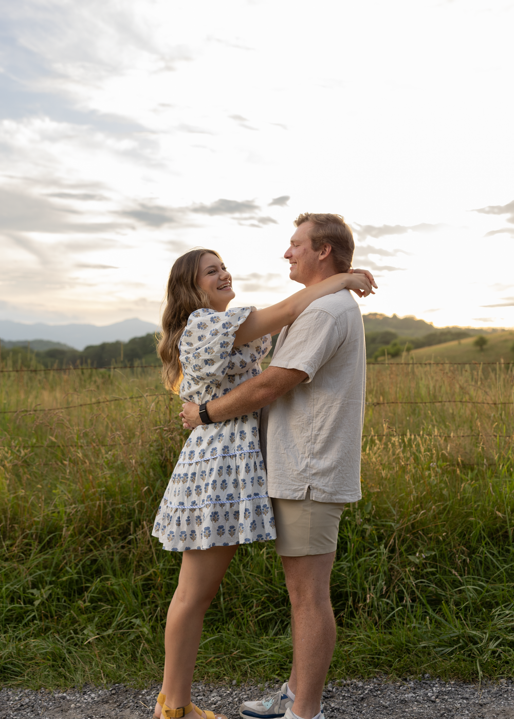 Couple embracing and smiling at each other on a path with a golden meadow and mountain sunset backdrop.