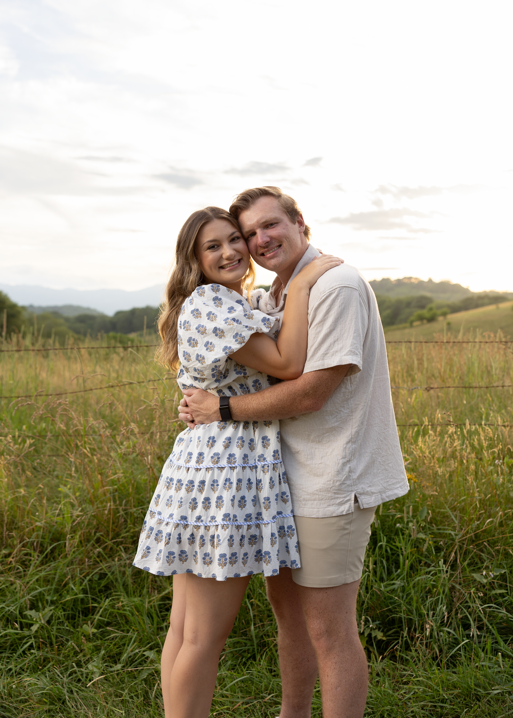 Couple hugging and smiling in a lush green field with rolling hills and a cloudy sky behind them.