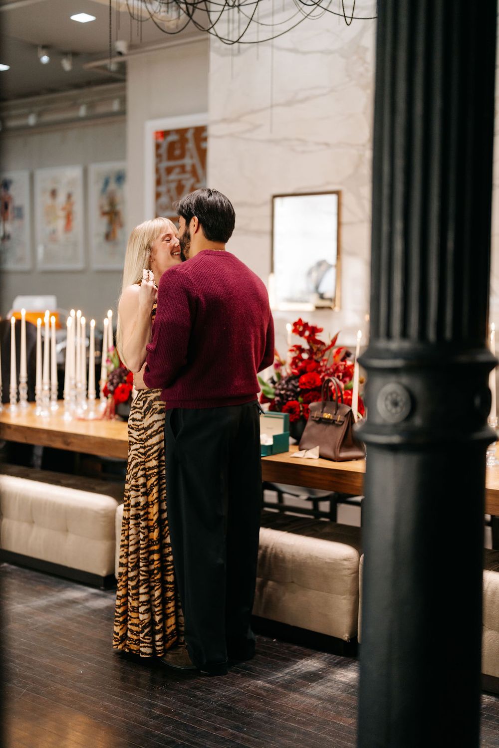 A couple stands close together at a candlelit table with red flowers and holiday decor in an elegant indoor setting.