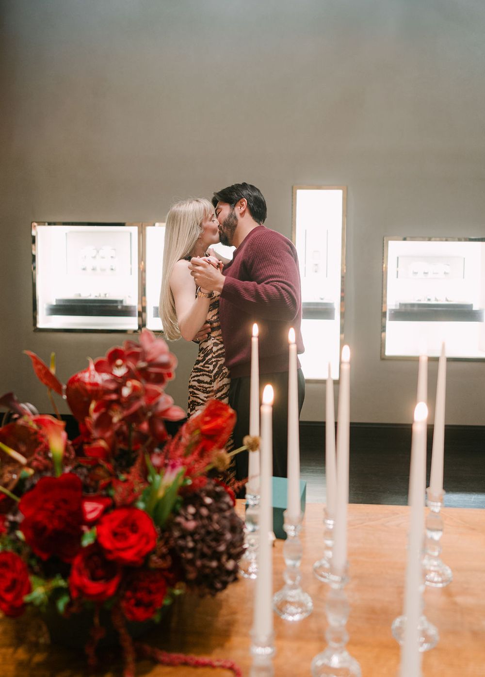 A romantic table setting with red floral centerpieces and tall white candles in glass holders.