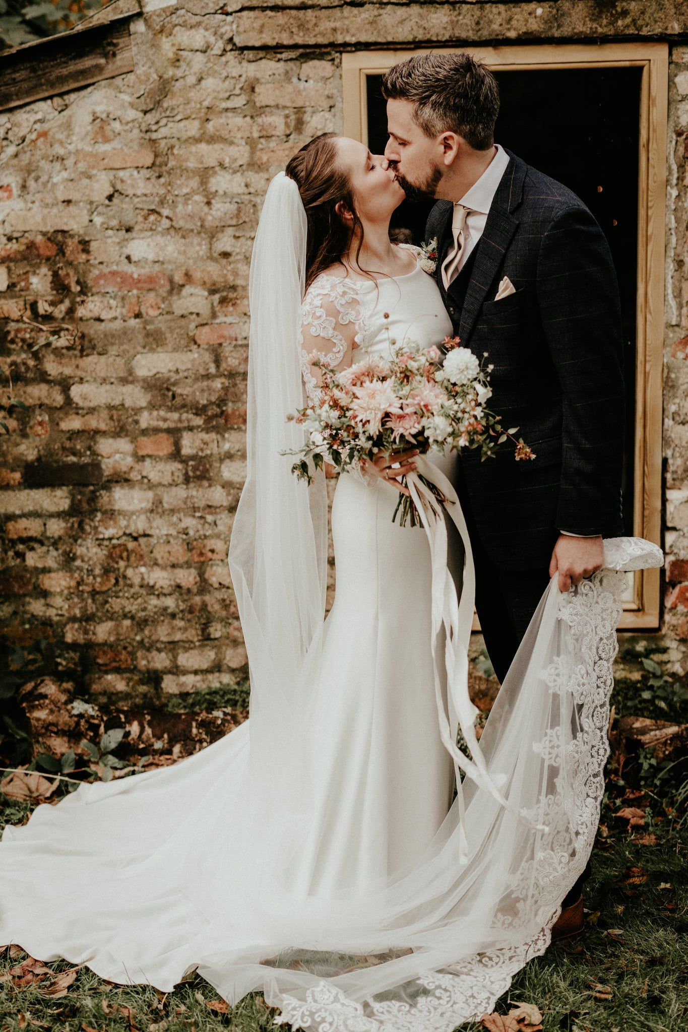 A romantic wedding couple shares a tender kiss against a rustic brick wall, with a flowing veil and delicate bouquet.