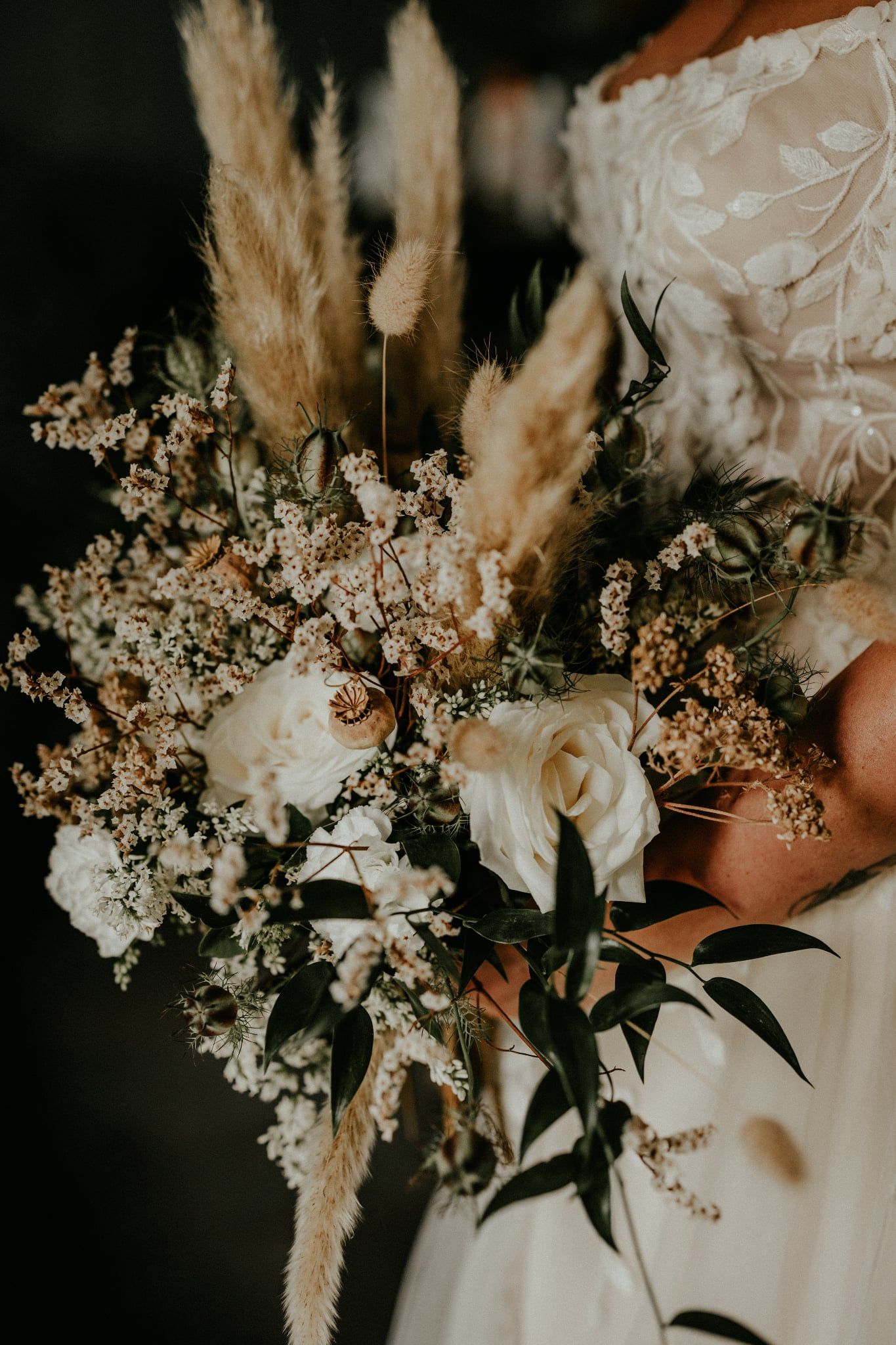 A collection of wedding bouquets featuring delicate white flowers, dried pampas grass, and dark green foliage in moody lighting.
