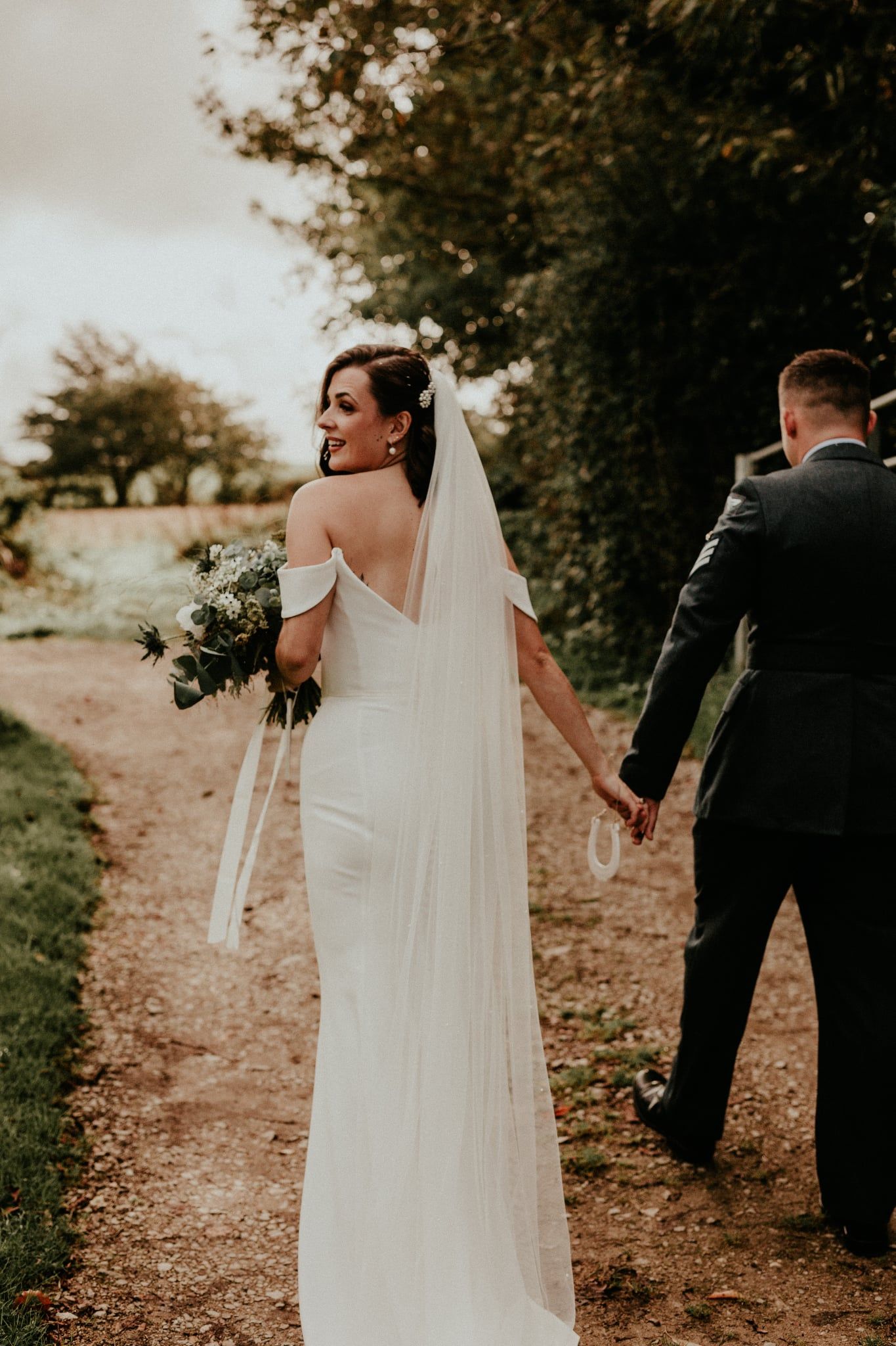 A couple in wedding attire walk along a rustic dirt path surrounded by trees during a moody autumn photoshoot.