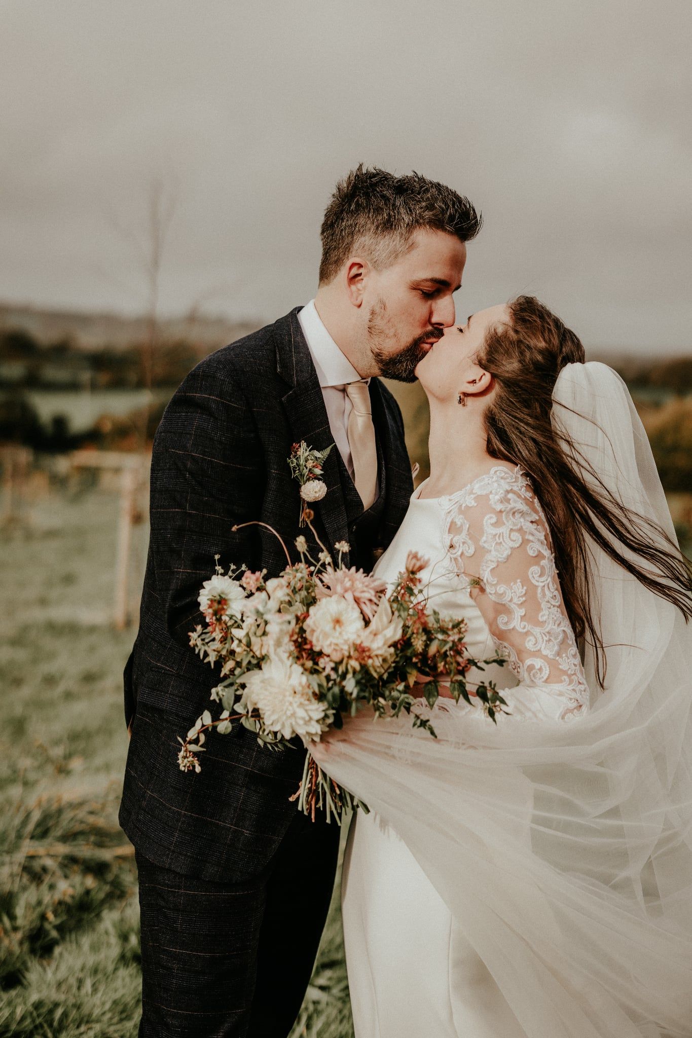 Romantic wedding couple shares a kiss in a field at sunset while holding a beautiful wildflower bouquet.
