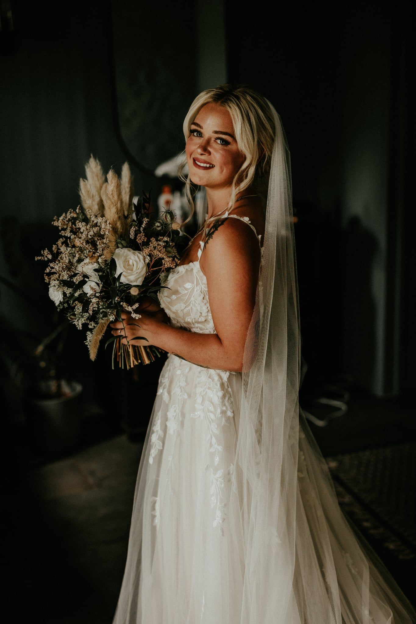 A bride in a flowing white lace wedding gown holds a neutral-toned bouquet against a dark background.