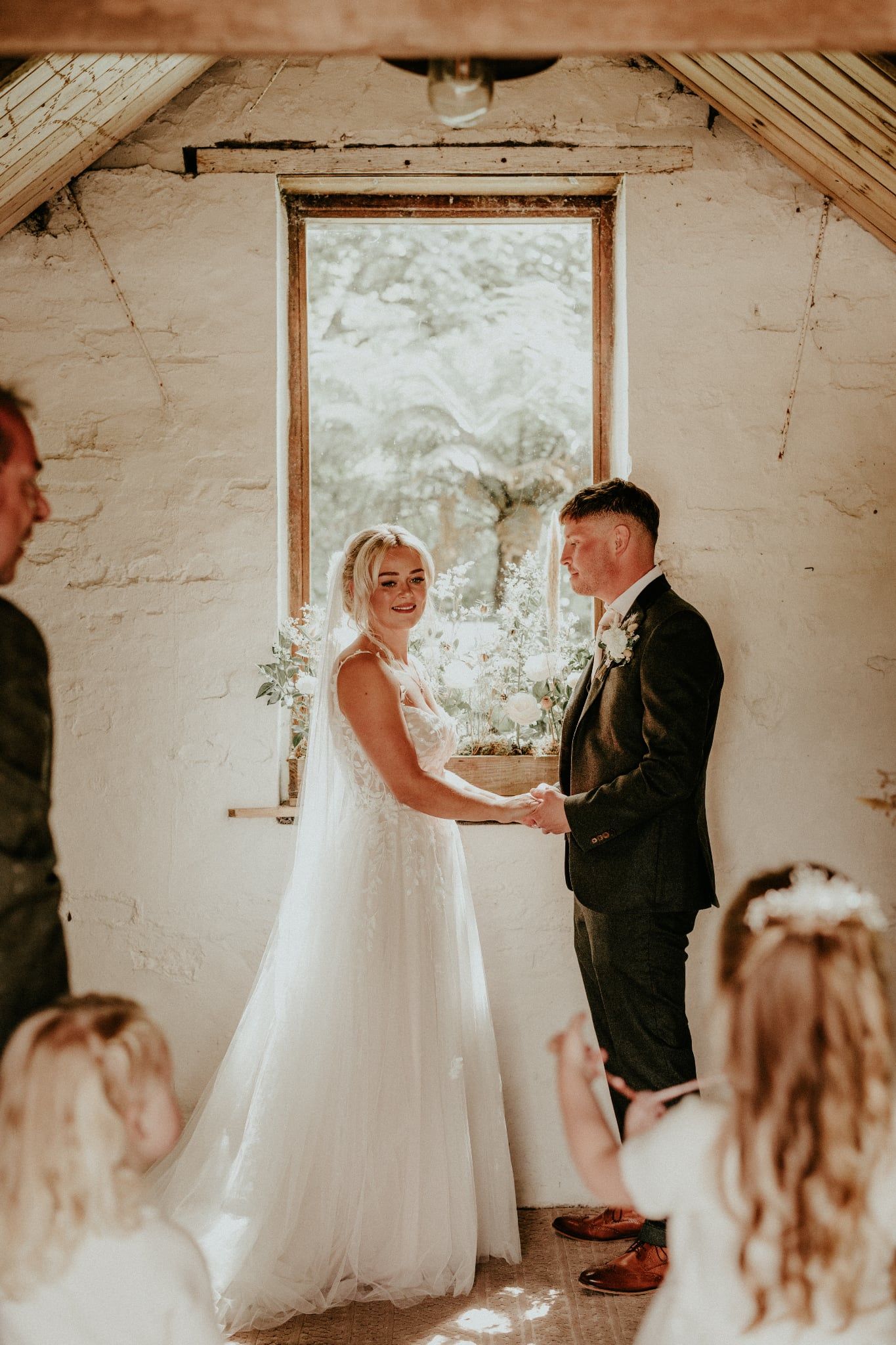 A romantic wedding ceremony takes place in a rustic white-walled room with natural light streaming through windows. Cornwall wedding photo