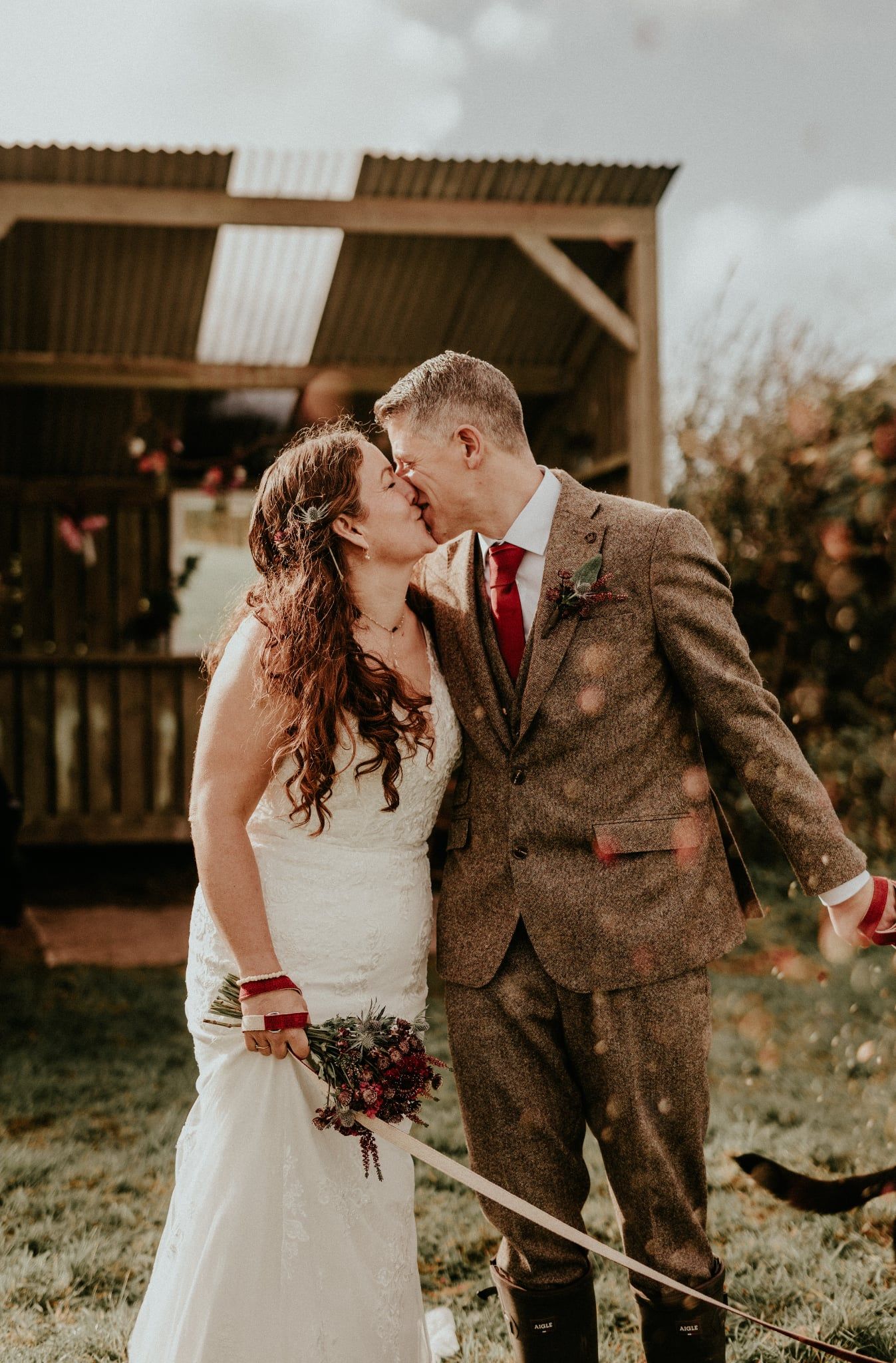 A romantic wedding couple sharing a kiss outdoors with falling autumn leaves and a rustic barn backdrop.