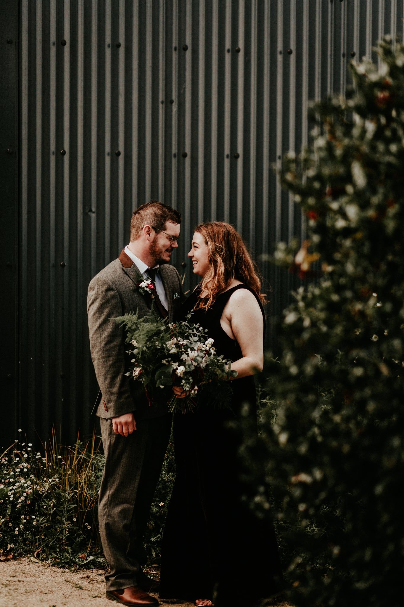 A couple shares an intimate moment in front of a dark corrugated wall with greenery in an outdoor setting.