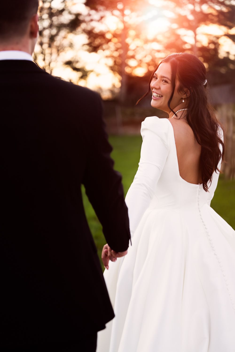 A joyful bride in a long-sleeved white gown looks back at her groom during sunset with warm golden light in background.