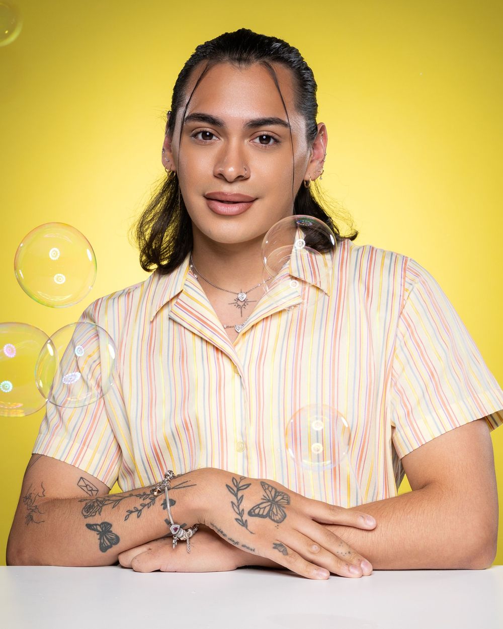 Someone wearing a pink striped shirt poses against a bright yellow background, showing off arm tattoos.