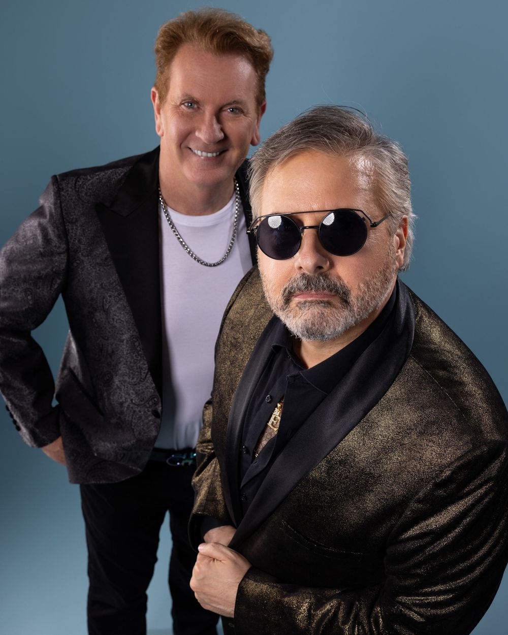 Two figures pose together in a series of studio photos against a blue background wearing black jackets and white shirts.