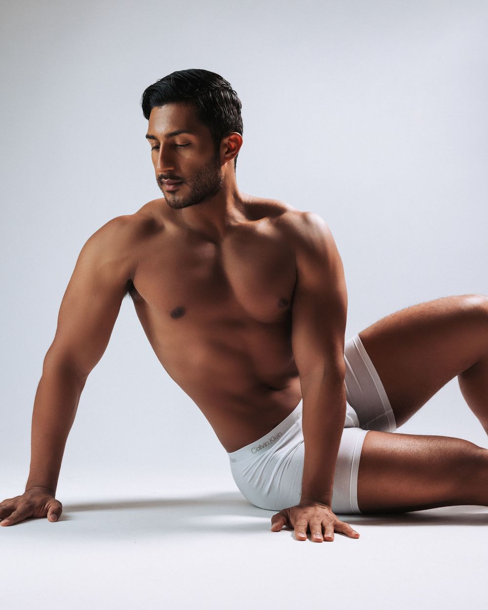 Artistic black and white photo of a muscular figure in white underwear reclining on light background.