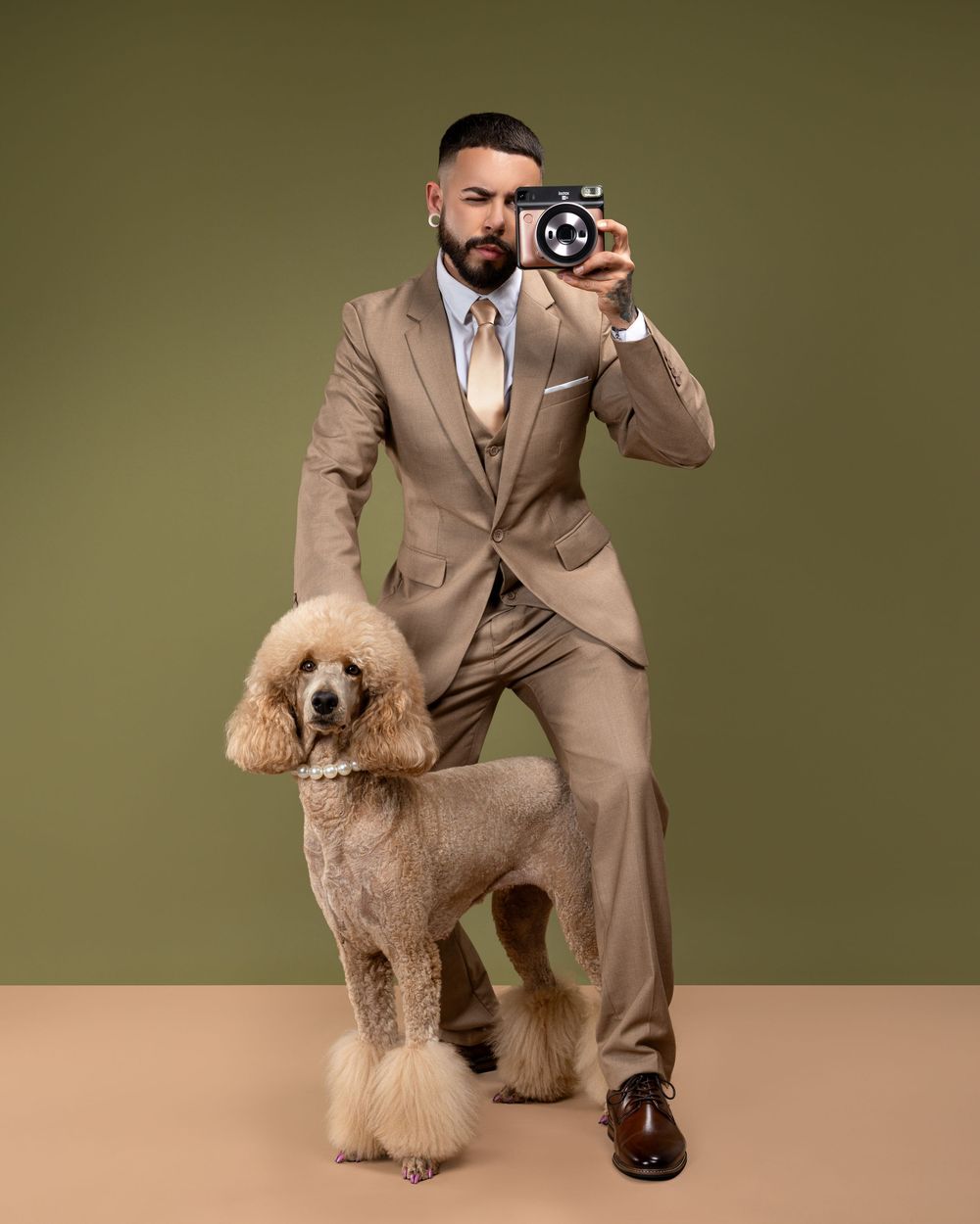 Professional photographer in tan suit takes pictures with a vintage camera while posing with a goldendoodle dog in studio.